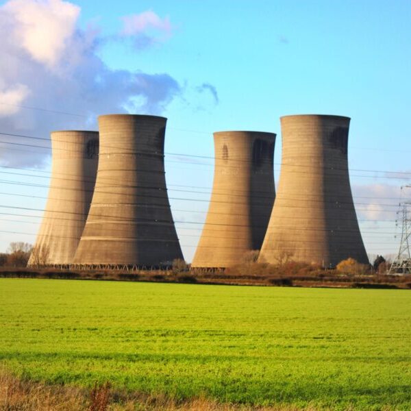 Four large cooling towers and power lines stand near a green field under a partly cloudy sky.